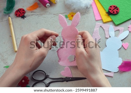Easter Bunny Felt Sewing Tutorial in Progress. Hands Stitching Pink Rabbit Shape with Needle, Surrounded by Colorful Materials, Flowers, Ladybugs, Threads, and Craft Tools for DIY Decoration