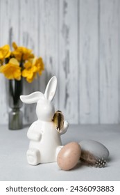 Easter bunny holding golden carrot, eggs on concrete table, bouquet of daffodils on background. Vertical greeting card or invitation, copy space. Happy Easter holidays