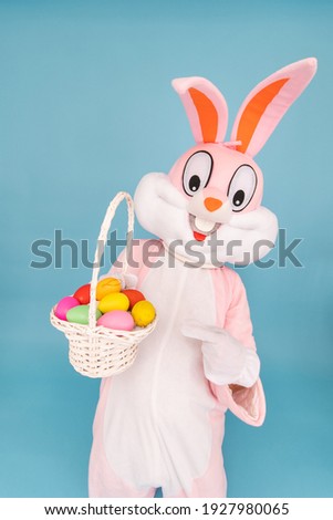 Easter bunny or rabbit or hare with basket of colored eggs, shows index finger, having fun, celebrates Happy easter. Easter rabbit isolated on blue background
