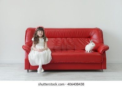 Easter Bunny Small And Cute With A Little Girl In A White Dress On The Red Couch In A Textural White Studio