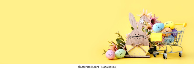 Easter Composition With Colorful Eggs In Shopping Cart, Wooden Bunny And Spring Flowers On Yellow Background. Banner. Copy Space