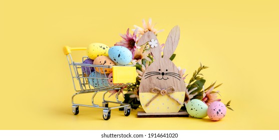 Easter Composition With Colorful Eggs In Shopping Cart, Wooden Bunny And Spring Flowers On Yellow Background. Banner.