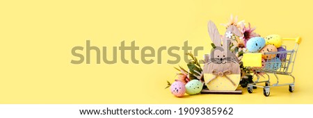 Easter composition with colorful eggs in shopping cart, wooden bunny and spring flowers on yellow background. Banner. Copy space