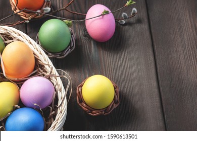 Easter composition - several colored eggs in a basket and on a dark wooden table with willow twigs, place for text, copy space
