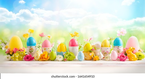 Easter concept with colorful decorated eggs in egg cups, bunny and flowers on white wooden table on spring nature background. Easter background with copy space.