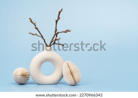 Easter concept. Ivory vase with willow branch and two paper craft Easter eggs on blue background monochrome