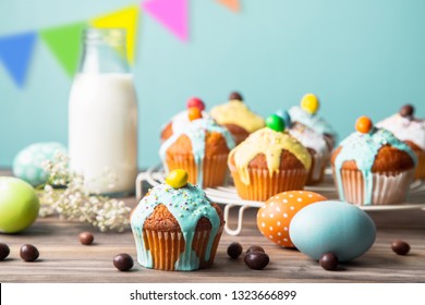 Easter cupcakes glazed with colorful sugar, sprinkles and chocolate with a bottle of milk and painted easter eggs  on a rustic wooden table against a turquoise background with pennants - Powered by Shutterstock