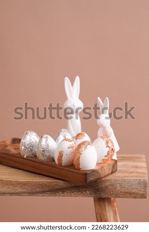 Easter decoration: eggs with silver and rose-gold metallic foil in a bamboos form on a wooden mediterranean style stool, white vase on beige colored seamless background, copy space