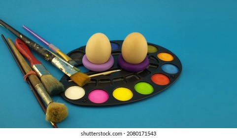 Easter Egg Decoration Concept With Paint Brushes And Set Of Watercolor Paints In Pallet. Studio Still Life On Blue Background