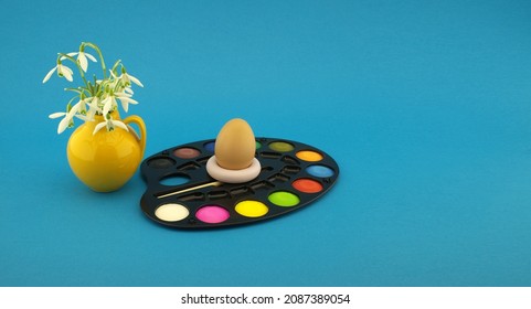 Easter Egg Decoration Concept With Set Of Watercolor Paints In Pallet, Next To A Yellow Jug With Fresh White Spring Flowers. Studio Still Life On Blue Background