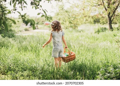 Easter egg hunt. Girl child Wearing Bunny Ears Running To Pick Up Egg In Garden. Easter tradition. Baby with basket full of colorful eggs. Back view