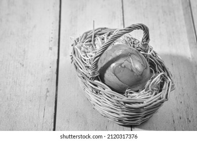 an easter egg in a woven wooden basket on a straw pad, on light wooden floorboards, black and white photo