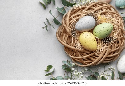 Easter eggs with pastel colors lying in wicker nest with straw and feathers, surrounded by flowers and eucalyptus leaves