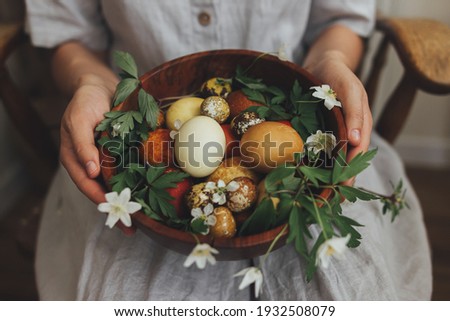Easter eggs and spring flowers in wooden bowl in hands on background of woman in rustic linen dress. Stylish easter and quail eggs in natural dye and spring blooms. Aesthetic holiday