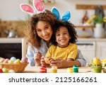 Easter Family traditions. Loving ethnic young mother teaching happy little kid soon to dye and decorate eggs with paints for Easter holidays while sitting together at kitchen table 