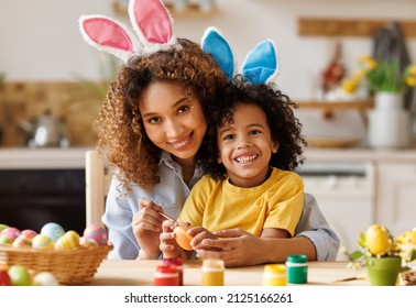 Easter Family traditions. Loving ethnic young mother teaching happy little kid soon to dye and decorate eggs with paints for Easter holidays while sitting together at kitchen table 