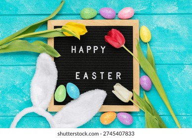 Easter flat lay composition featuring a black letter board with text Happy Easter decorated with colorful glitter Easter eggs, white bunny ears and fresh tulips on a rustic blue wooden background.