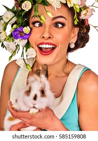Easter girl holding bunny. Woman with holiday spring flowers hairstyle and make up with fake eyelashes. Adults at the festival. Female makes squint eye for fun. She has strabismus on white background.