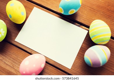 easter, holidays, tradition and object concept - close up of colored easter eggs and blank white paper card on wooden surface with copy space