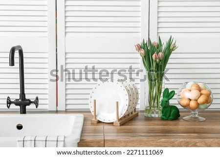 Easter kitchen interior with pastel eggs in glass bowl, pink tulips in vase, green bunny figurine, white plates on wooden countertop and farmhouse sink