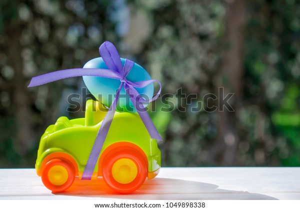 Easter light\
green car with a blue egg tied with a purple ribbon rides on the\
table against the background of\
greenery