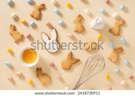 Easter pattern with bunnies shaped cookies, eggs, sweet chocolate eggs, themed cookie cutters and whisk on beige background. Festive food and snacks. Greeting card. Flat lay style.