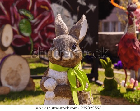 Easter in spring season with a cute bunny decoration with a green scarf, and colorful blurred background includes trees, eggs, wooden log, window, green grass in Mosonmagyaróvár, Hungary, Europe.