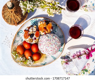 Easter Still Life In Garden - Traditional Food On White Rustic Tablecloth, Outdoors. Traditional Bread Paska Or Kulich, Colored Eggs, Red Sweet Wine And Bouquet Of  Flowering Cherry Sprigs, Top View 