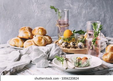 Easter table setting with colored orange eggs, hot cross buns, green branches decorated, empty white plate with cutlery, glass of lemonade drink over white plank wooden table with textile tablecloth.