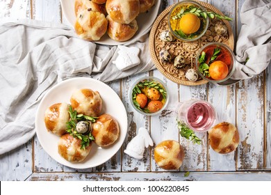 Easter table setting with colored orange eggs, hot cross buns, green branches decorated, empty plate, cutlery, glass of lemonade drink over white plank wooden table with textile tablecloth. Flat lay