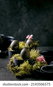 Easter table setting with empty black ceramic plates and mugs, decorated by forest moss, black colored easter eggs and pink spring flowers on wooden table. Dark stylish holidays.
