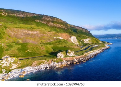 The Eastern Coast Of Northern Ireland And Antrim Coast Road, A.k.a. Giant's Causeway Coastal Route With A Car. Aerial View At Sunrise. Carnlough In The Background