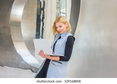 Eastern European Business Woman Working At High Tech Firm In New York, Wearing Long Vest, Black Undershirt, Leaning Back On Metal Structure, Working On Laptop Computer. 
