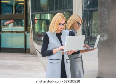 Eastern European Business Woman Working At High Tech Firm In New York, Wearing Long Vest, Black Undershirt, Glasses, Standing By Metal Mirror, Working On Laptop Computer.
