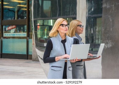 Eastern European Business Woman Working At High Tech Firm In New York, Wearing Long Vest, Black Undershirt, Glasses, Standing By Metal Mirror, Working On Laptop Computer.
