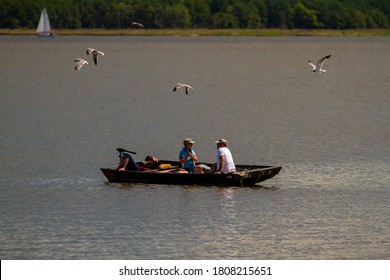 Eastern Neck Island, 08/30/2020: Three People Are Relaxing On A Small Wooden Fishing Boat. One Is Sleeping The Other Two Are Talking. A Flock Of Water Birds Are Flying Over Them Trying To Steal Food.