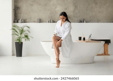 Eastern Woman In Bathrobe Moisturizing Smooth Legs With Moisturizer Cream Sitting On Bathtub In Modern Bathroom Indoors After Morning Shower. Beauty, Skincare And Pampering Concept, Copy Space