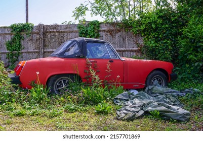 Eastleigh, Hampshire, England - 05.17.2022 :  A neglected MG Midget car on scrubland. A red two seater sports car built in the 1970's with soft top roof. Classic old English sports car.