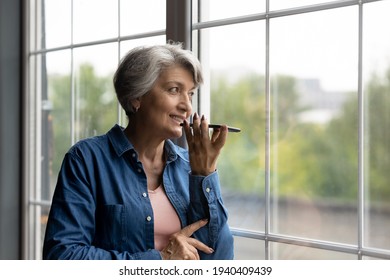 Easy communication. Confident aged hispanic female look at large window hold modern smartphone dictate audio message. Smiling senior latina woman give vocal command to digital ai assistant. Copy space - Powered by Shutterstock