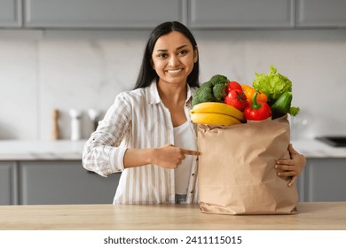 Easy Grocery Shopping. Young Indian Woman Pointing At Paper Bag With Fresh Vegetables And Fruits, Happy Lady Enjoying Organic Healthy Food, Standing In Cozy Kitchen At Home, Copy Space - Powered by Shutterstock