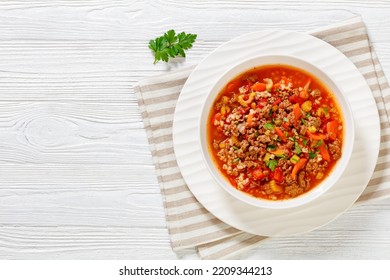 Easy hot and hearty Hamburger Soup with barley and vegetables in white bowl on white wood table, horizontal view from above, flat lay, free space