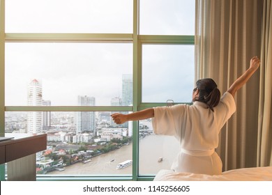 Easy lifestyle Asian woman waking up from good sleep in weekend morning taking some rest, relaxing in comfort bedroom at hotel window, having happy lazy day enjoying work-life quality balance concept