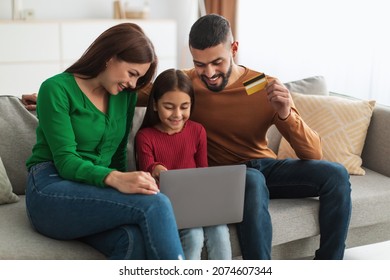 Easy Payment And Cashback Concept. Portrait Of Smiling Middle Eastern Family Doing Online Shopping At Home Using Laptop Pc, Man Holding Debit Credit Card In Hand, Three People Sitting On Couch At Home