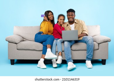 Easy Payment And Cashback Concept. Portrait of smiling black family doing online shopping using laptop pc, woman holding debit credit card in hand, three people sitting on couch at blue studio wall