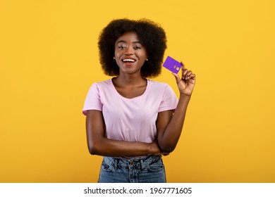 Easy Payment Concept. Happy Young Black Woman Holding Debit Credit Card In Hand, Smiling And Showing It To Camera, Posing Isolated Over Yellow Studio Background