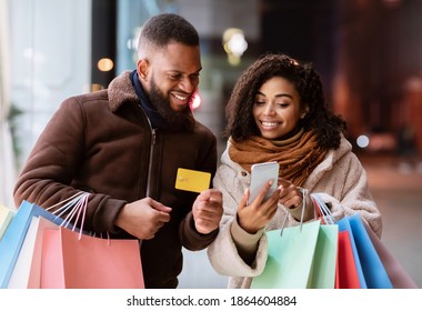 Easy Payment Concept. Portrait of happy african american man and woman using mobile phone, holding credit card and shopping bags, standing outdoors in the evening. Retail And Purchase