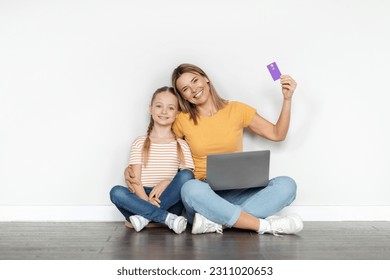 Easy Payments. Cute Little Girl And Her Happy Mother With Laptop And Credit Card Sitting On Floor Near White Wall, Smiling Millennial Woman And And Female Child Enjoying E-Commerce, Copy Space - Shutterstock ID 2311020653