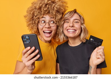 Einfache Zahlungen. Happy junge Frauen machen Internet-Shopping mit Mobiltelefon und Bankkarte kaufen Dinge online Lächeln breit gekleidet in Casual-t-Shirt einzeln auf lebendig gelbem Hintergrund