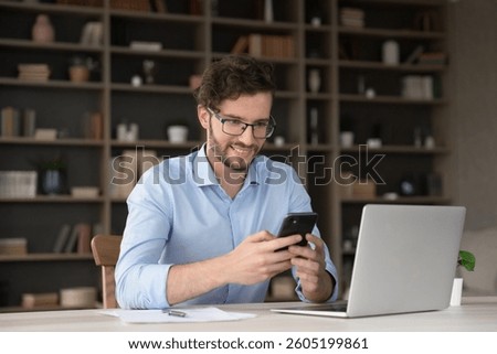 Easy and productive multitasking. Smiling young man remote working employee sit at workplace by notebook interact with phone app to send text message to colleague check social media during work break