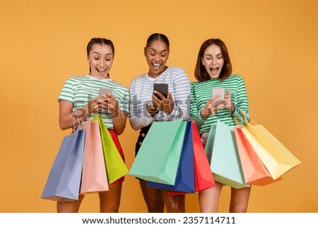 Easy shopping, black friday deal, sale season, ecommerce. Amazed happy three young multiethnic ladies shopping online, using smartphones, holding colorful shopping bags, orange background